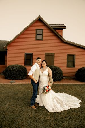 Wedding couple poses in front of rustic red farmhouse with flowing lace train.