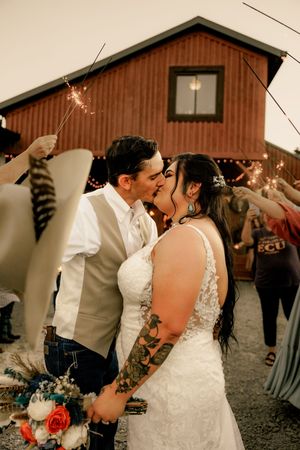 Romantic sparkler kiss moment between newlyweds outside rustic barn venue at dusk.