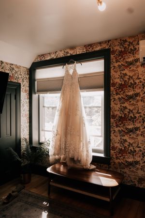 A delicate white lace wedding dress hangs in a room with floral wallpaper and natural lighting.