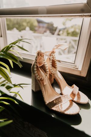 Elegant sparkly wedding heels sit on a windowsill with green plants in soft natural light.