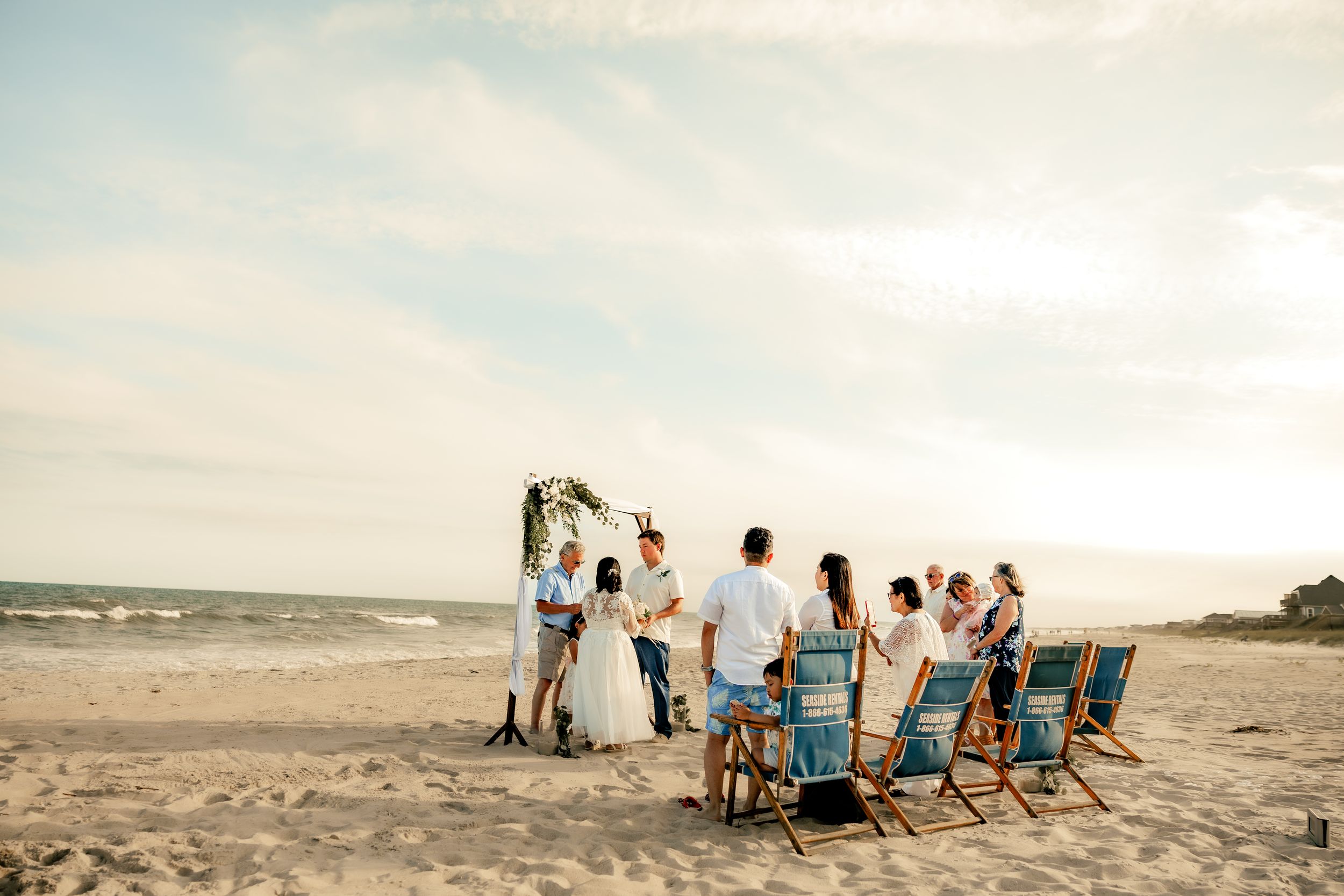 Small beach wedding ceremony with guests seated in blue chairs facing couple at floral arch by the ocean.