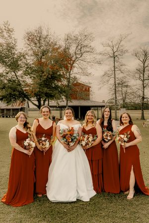 Bridesmaids in burgundy dresses and bride in white pose together on grass during autumn wedding.