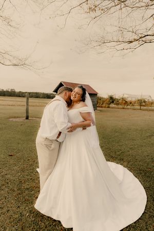 A joyful couple in white wedding attire embraces and spins together in a scenic outdoor field at sunset.
