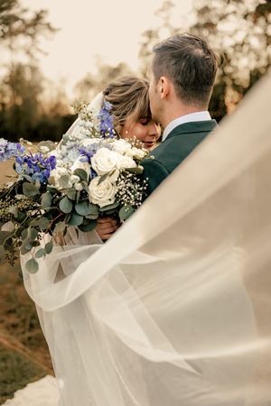 Ethereal wedding photos with flowing veil and blue and white floral bouquet in natural outdoor setting.