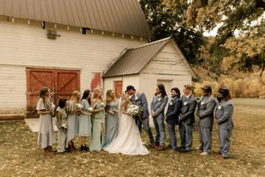 Wedding party poses in front of a rustic white barn with bridesmaids in sage and groomsmen in grey.