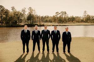 Wedding party members in formal attire create silhouettes against sunset lake backdrop.