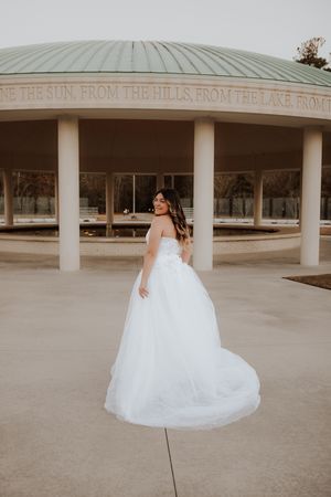 Bride in flowing white gown poses beneath circular pavilion during wedding photoshoot.