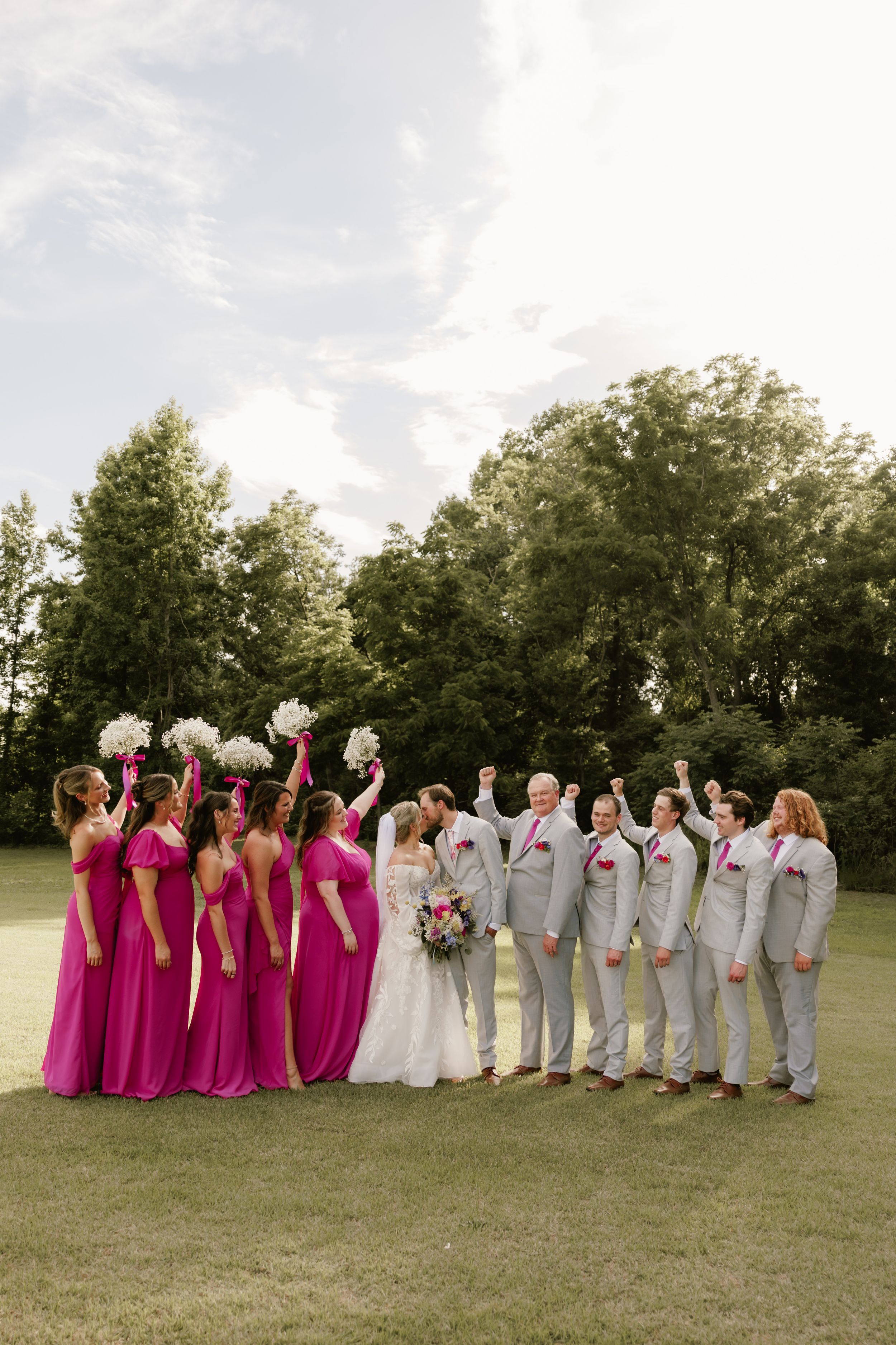 Wedding party celebrating outdoors, bridesmaids in fuchsia gowns and groomsmen in gray suits cheering with bride and groom.