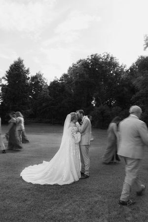 Black and white wedding photo of bride and groom sharing a kiss outdoors while guests mill around them.