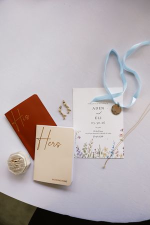 Wedding flat lay with vow books, floral invitation for Aden and Eli, blue ribbon, gold necklace, and earrings on white table.