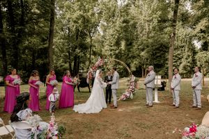 Outdoor wedding ceremony with pink bridesmaids, floral circular arch, bride and groom exchanging vows in a lush green forest.