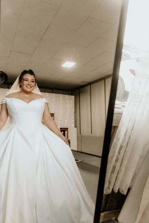 Beautiful off-shoulder white wedding gown with full ballgown skirt shown in mirror reflection with string lights in background.