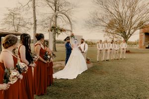 A romantic outdoor wedding ceremony with bridesmaids in burgundy dresses standing in a row on a grassy field.