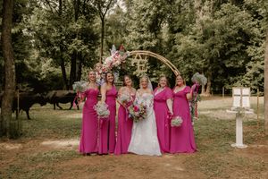 Bride in white gown poses with five bridesmaids in pink dresses holding bouquets under floral arch outdoors.