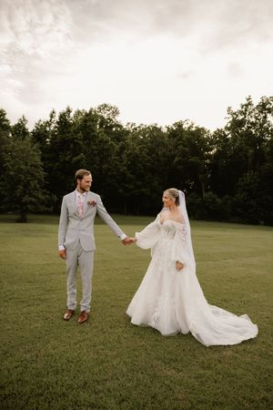 A bride in a flowing white gown and groom in a gray suit hold hands on a lush green lawn with trees behind them.