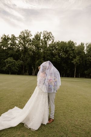 Bride and groom sharing a kiss under a flowing veil on a lush green lawn with trees in the background.