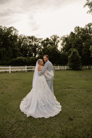 Bride and groom embrace in a lush green field, her lace gown trailing elegantly, with a white fence and trees behind.