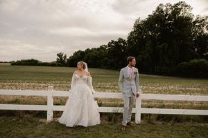 Bride in lace gown and groom in gray suit stand by white fence at rustic farm wedding venue.