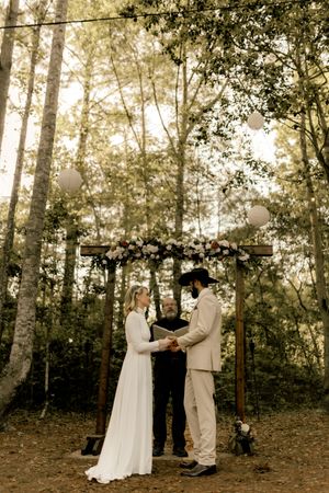 Bride and groom exchange vows under a floral wooden arch in a sunlit forest wedding ceremon in Wilmington, NC.