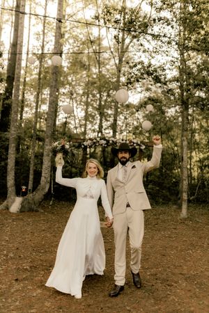 Joyful newlyweds celebrate in a forest wedding venue, bride in white gown and groom in beige suit raising arms.