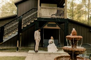 Bride and groom pose outside a dark green barn venue with a stone fountain and wooded backdrop.