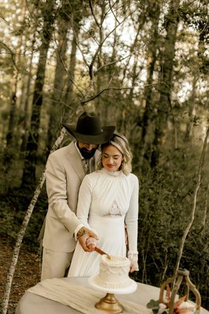 Bride and groom cutting wedding cake outdoors in forest, groom wearing cowboy hat and tan suit.