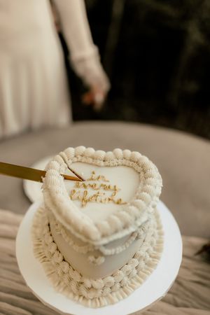 Heart-shaped white wedding cake with gold script and ruffled frosting detail, held by bride in white dress.