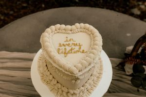 Heart-shaped white wedding cake with gold 'in every lifetime' script, decorated with rosette piping on a white stand.