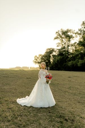 Bride in white lace gown holding red bouquet stands in golden field at sunset with trees in background.