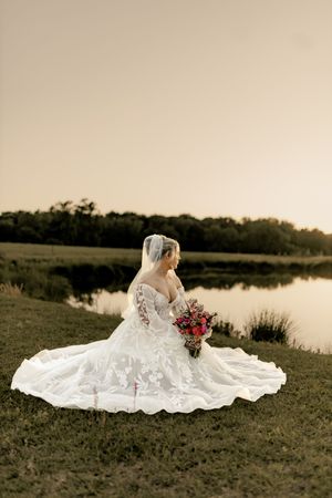 Bride in white lace gown with long train and veil holds pink bouquet by serene lake at sunset in Wilmington.