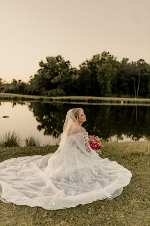 Bride in a flowing white lace gown with veil sits by a reflective lake at sunset, holding a vibrant bouquet at Wrightsville manor in Wilmin.