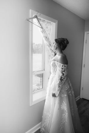 Bride in lace off-shoulder wedding gown holding dress on hanger near window, black and white photo.