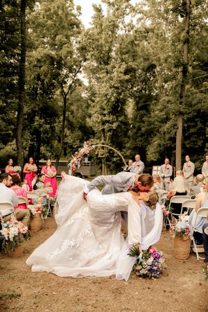 Groom dips bride for a kiss at outdoor woodland wedding ceremony with guests seated on white chairs.