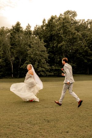 Bride in white gown twirls as groom chases her across green field at sunset with trees in background.