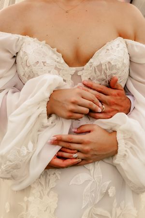 Bride in lace off-shoulder wedding gown with groom's arms wrapped around her, both wearing wedding rings.