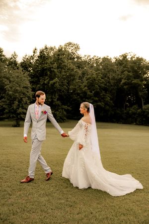 Bride in white gown and groom in gray suit hold hands walking across green lawn at golden hour wedding.