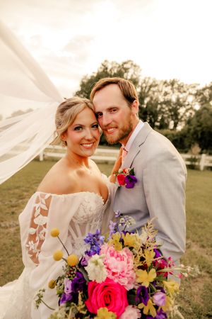 Smiling bride and groom pose outdoors with colorful bouquet, veil flowing in breeze at Wilmington, NC wedding venue.