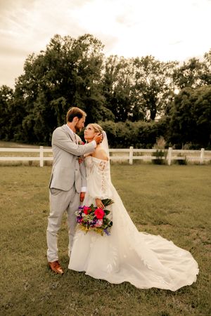 Bride and groom share romantic embrace on green lawn at sunset, she holds colorful bouquet near white fence.