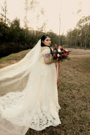 A bride in a flowing white lace wedding gown poses with a deep red bouquet in a rustic outdoor setting at sunset.
