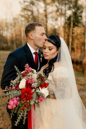A romantic wedding couple poses in an autumn forest setting with vibrant red and pink floral bouquet details.