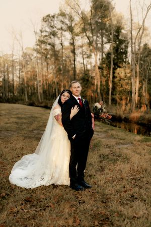 A couple poses for their wedding photos in an autumn forest setting with a long white veil and formal attire.