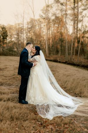 A couple shares a romantic moment in a wooded setting at sunset, with the bride's long flowing veil trailing behind her.