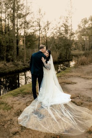 A couple in wedding attire embraces on a wooded path as the bride's long veil trails dramatically behind them.