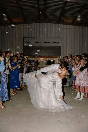 Groom dips bride for a romantic kiss surrounded by bubbles and cheering guests at elegant barn wedding reception.