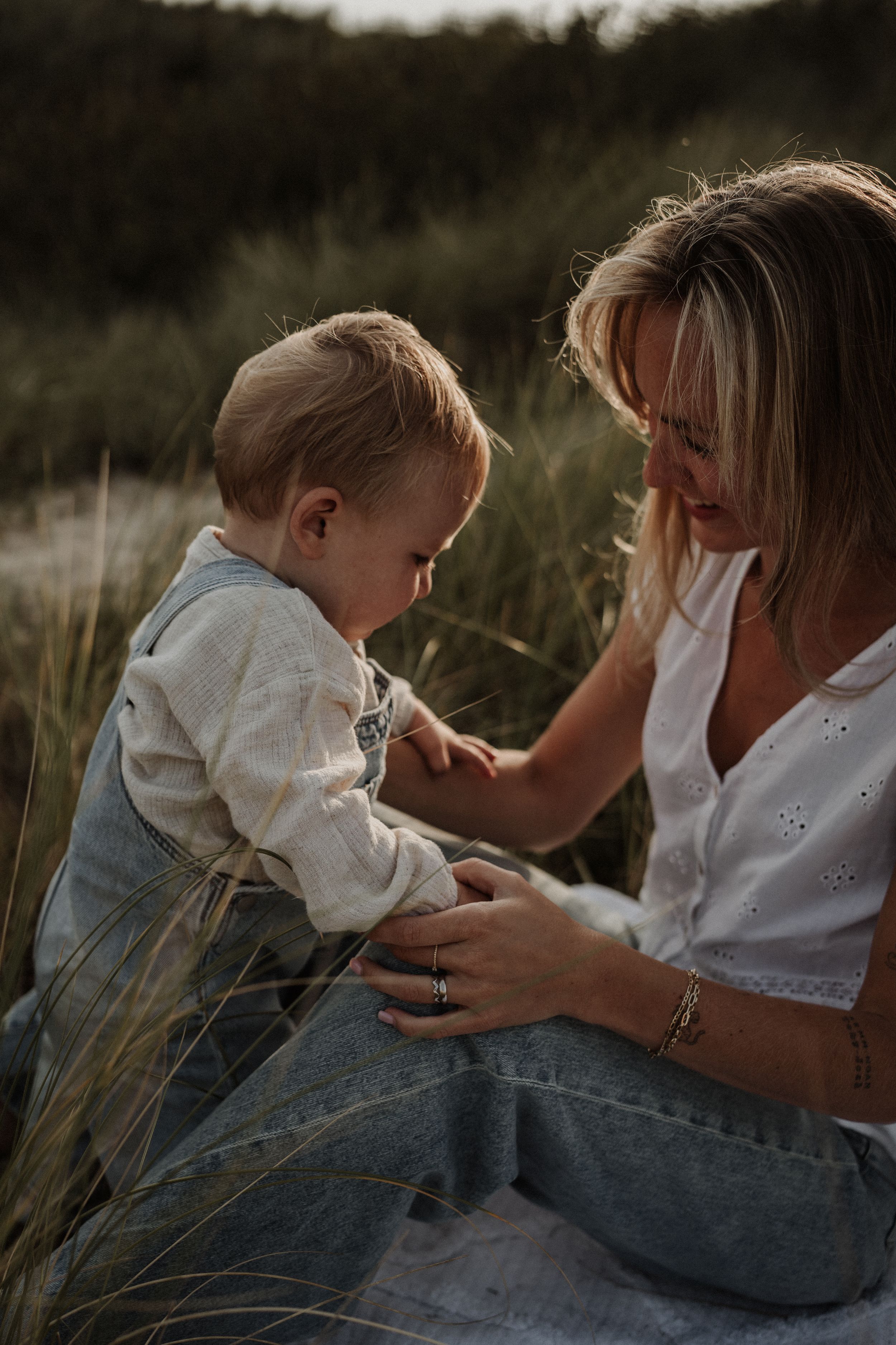 A tender moment captured outdoors as a parent and young child share a connection while sitting in tall grass at sunset.