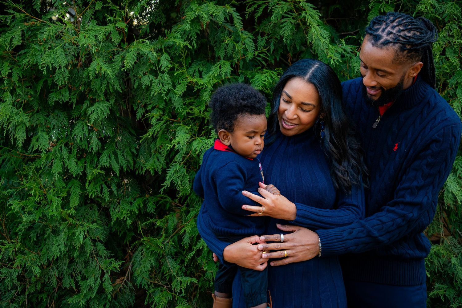Family wearing matching navy sweaters share a warm embrace against evergreen trees.