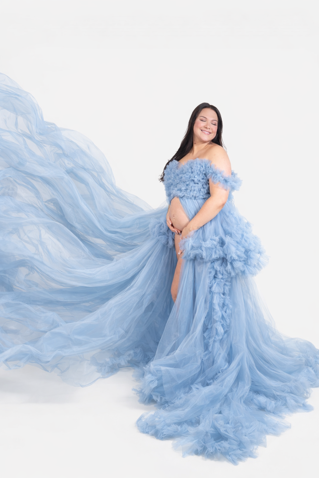 Pregnant woman in flowing blue ruffle tulle gown posing against white studio background.