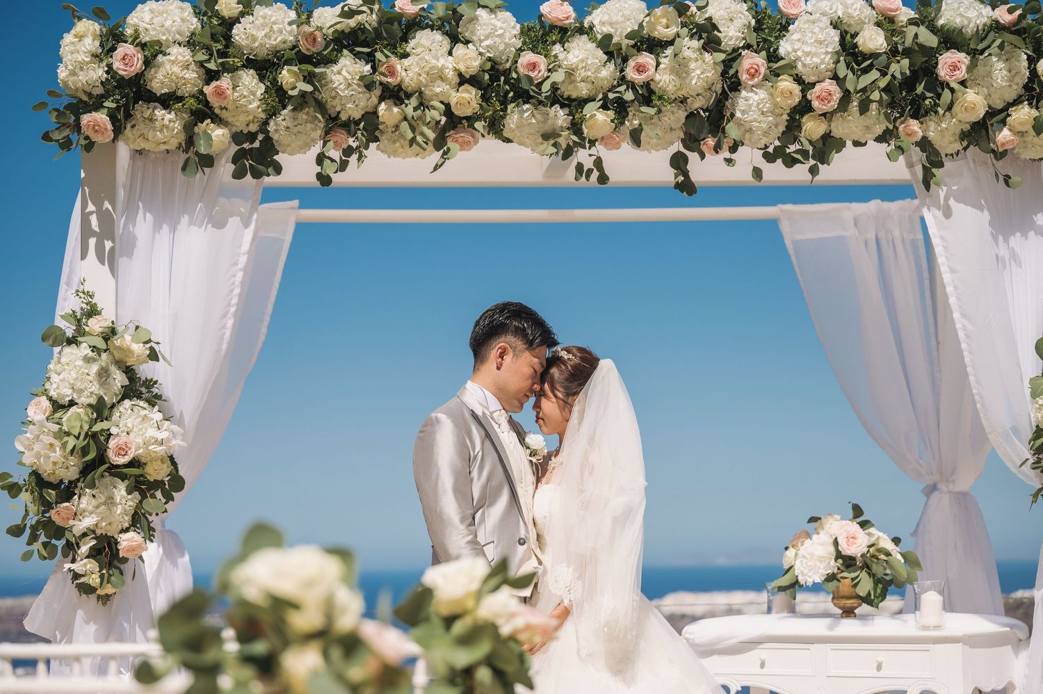 A romantic beach wedding ceremony under a white draped arch adorned with cream roses against a blue ocean backdrop.