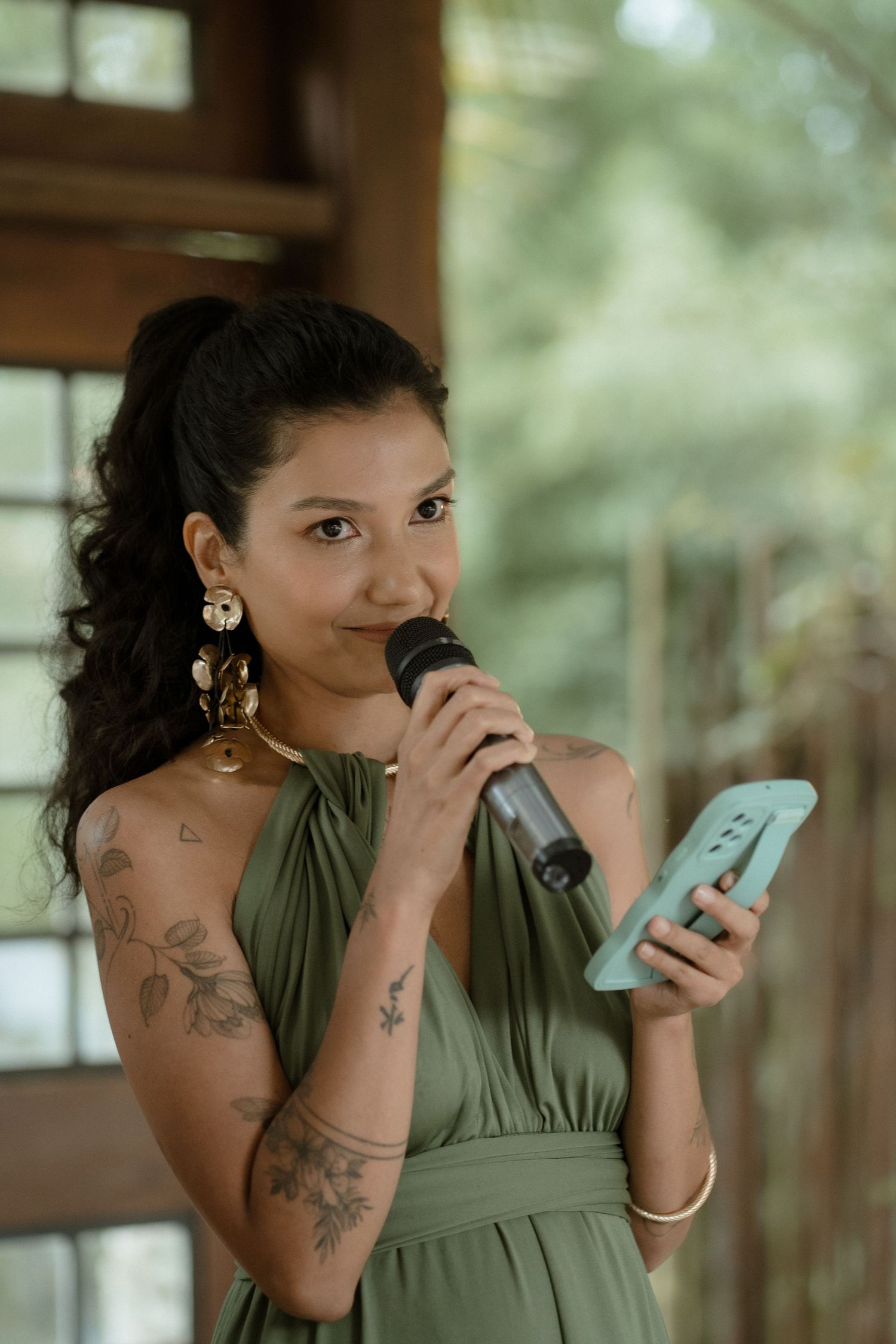 Someone in a sage green dress gives a speech while holding a phone and microphone at an outdoor event.