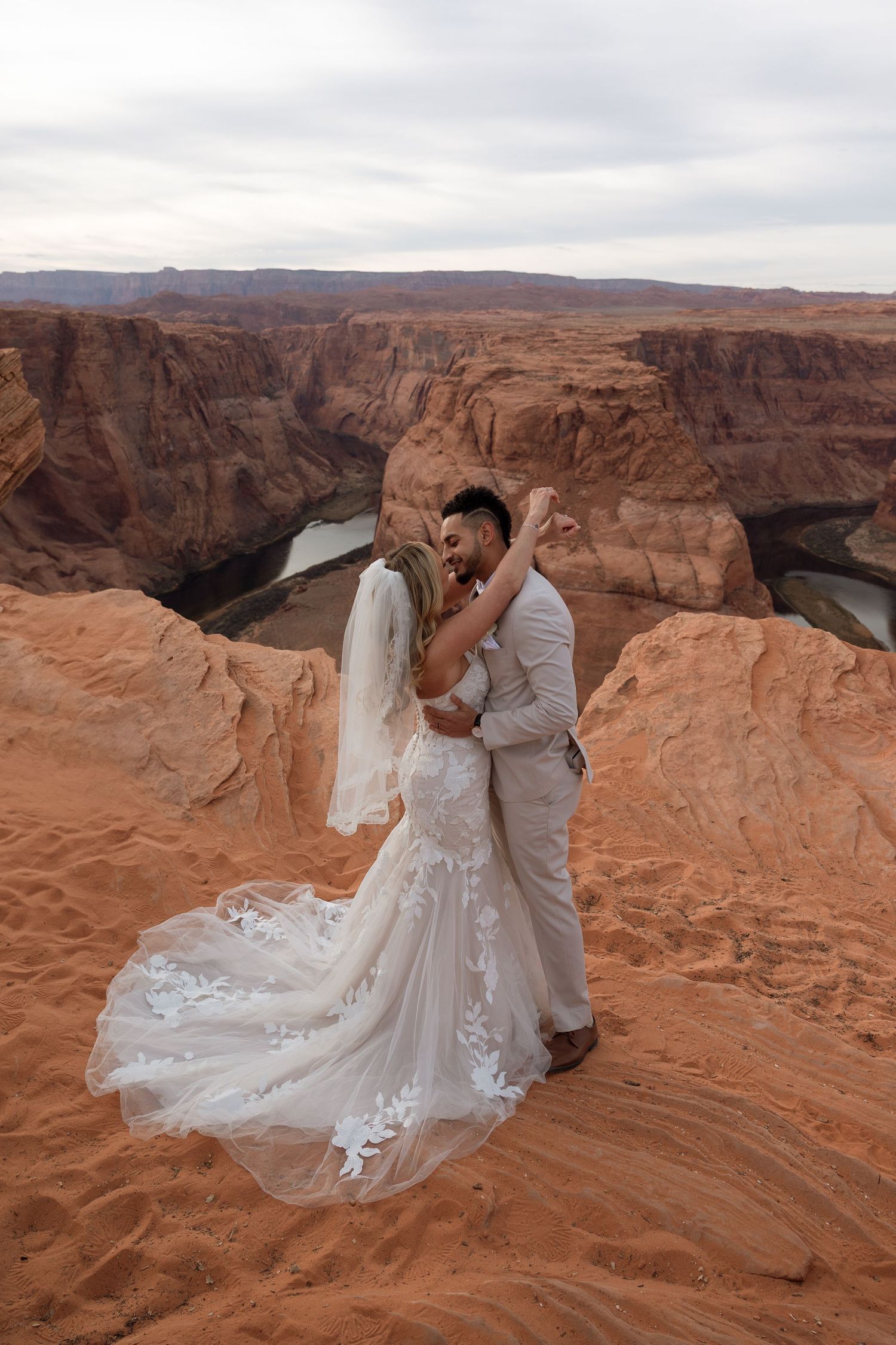 A wedding couple embraces on red sandstone cliffs overlooking a dramatic desert canyon landscape.